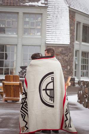 Two people wrapped in a blanket with a symbol, standing outdoors in front of a building with snow on the ground.