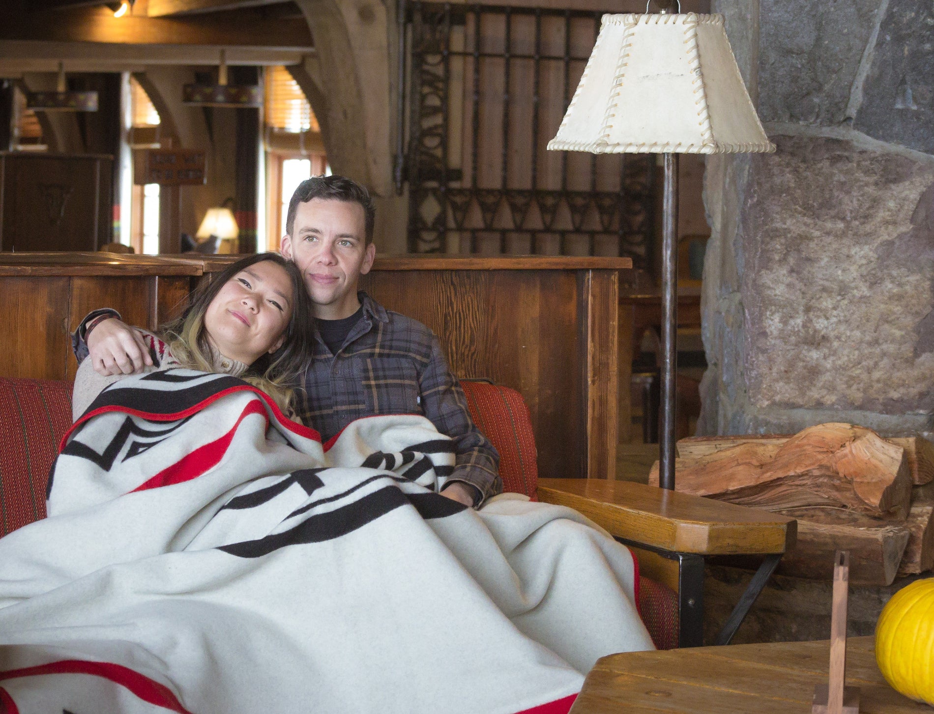 Two people sitting together under a blanket in a cozy room with wooden furniture and stone wall.