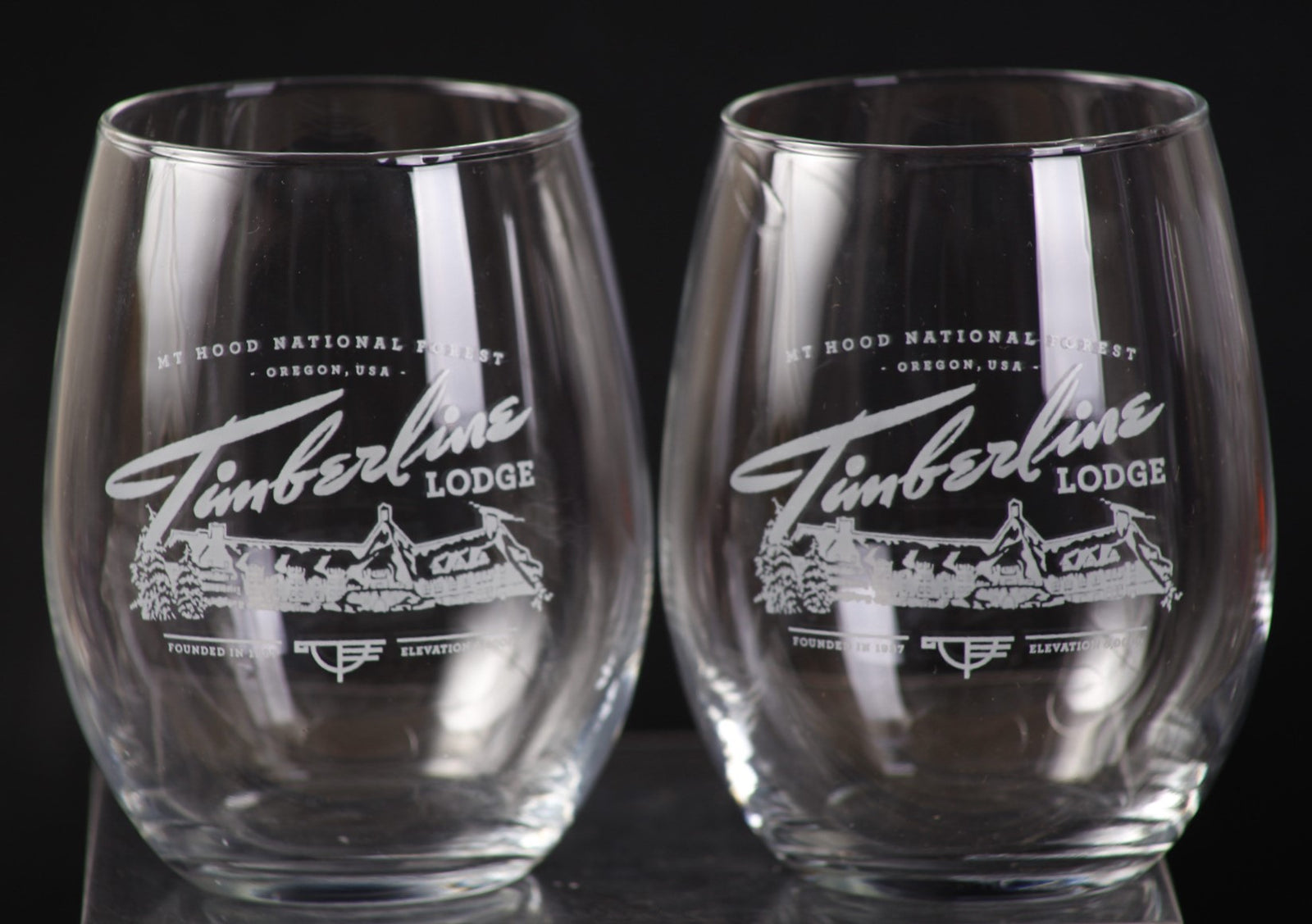 Two clear stemless wine glasses with engraved text and logo on a dark background